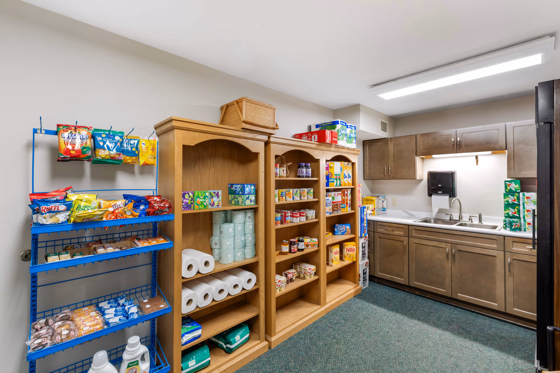 Small pantry/kitchen area with wooden shelves stocked with snacks and paper goods next to a countertop, sink, and cabinets.