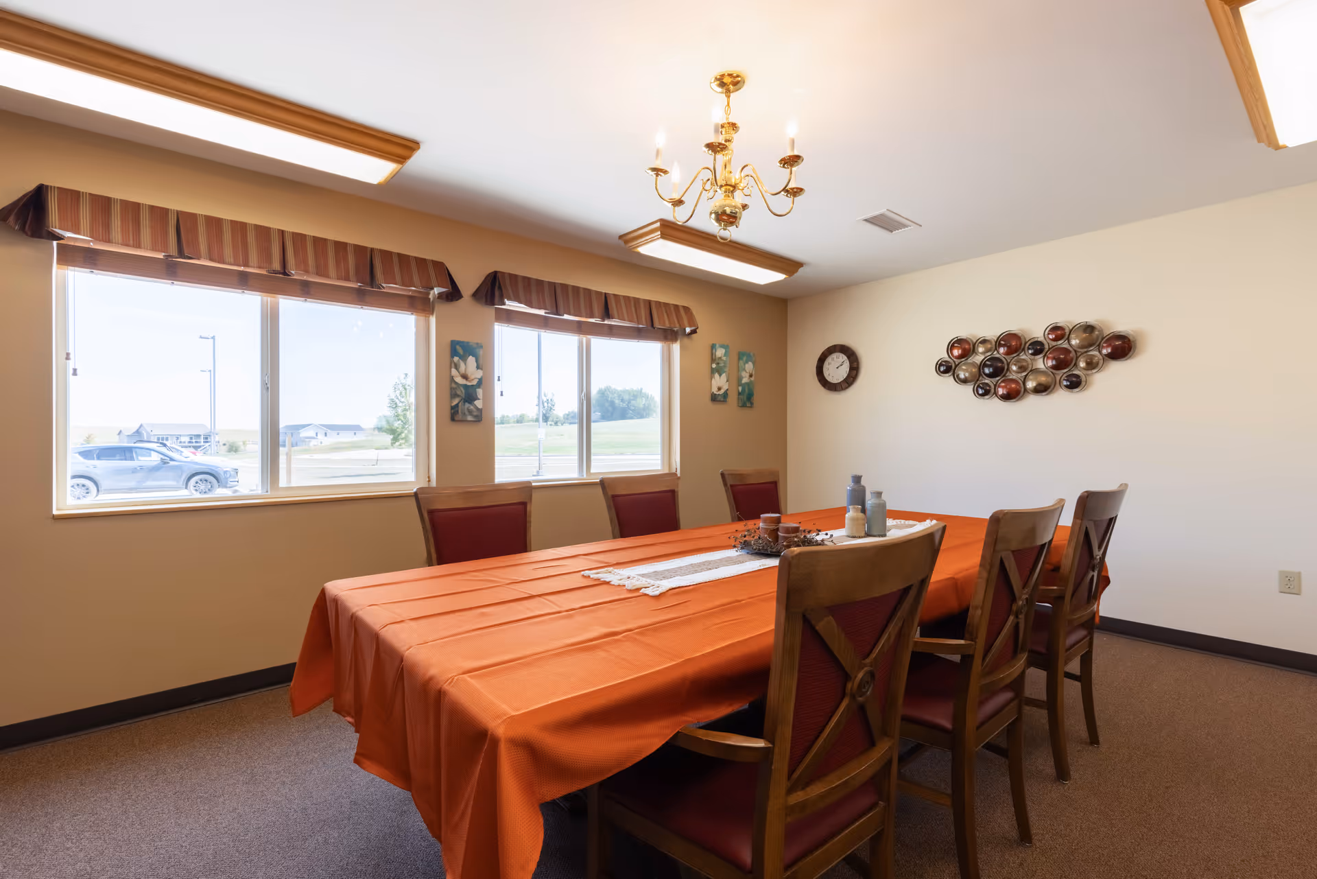 A dining room with a long rectangular table covered with an orange tablecloth and a white runner. Six wooden chairs with red cushions surround the table. The room has three large windows with striped valances, letting in natural light. On the wall, there is a round clock and a decorative metal wall art piece. The ceiling has a gold chandelier and two rectangular fluorescent lights.