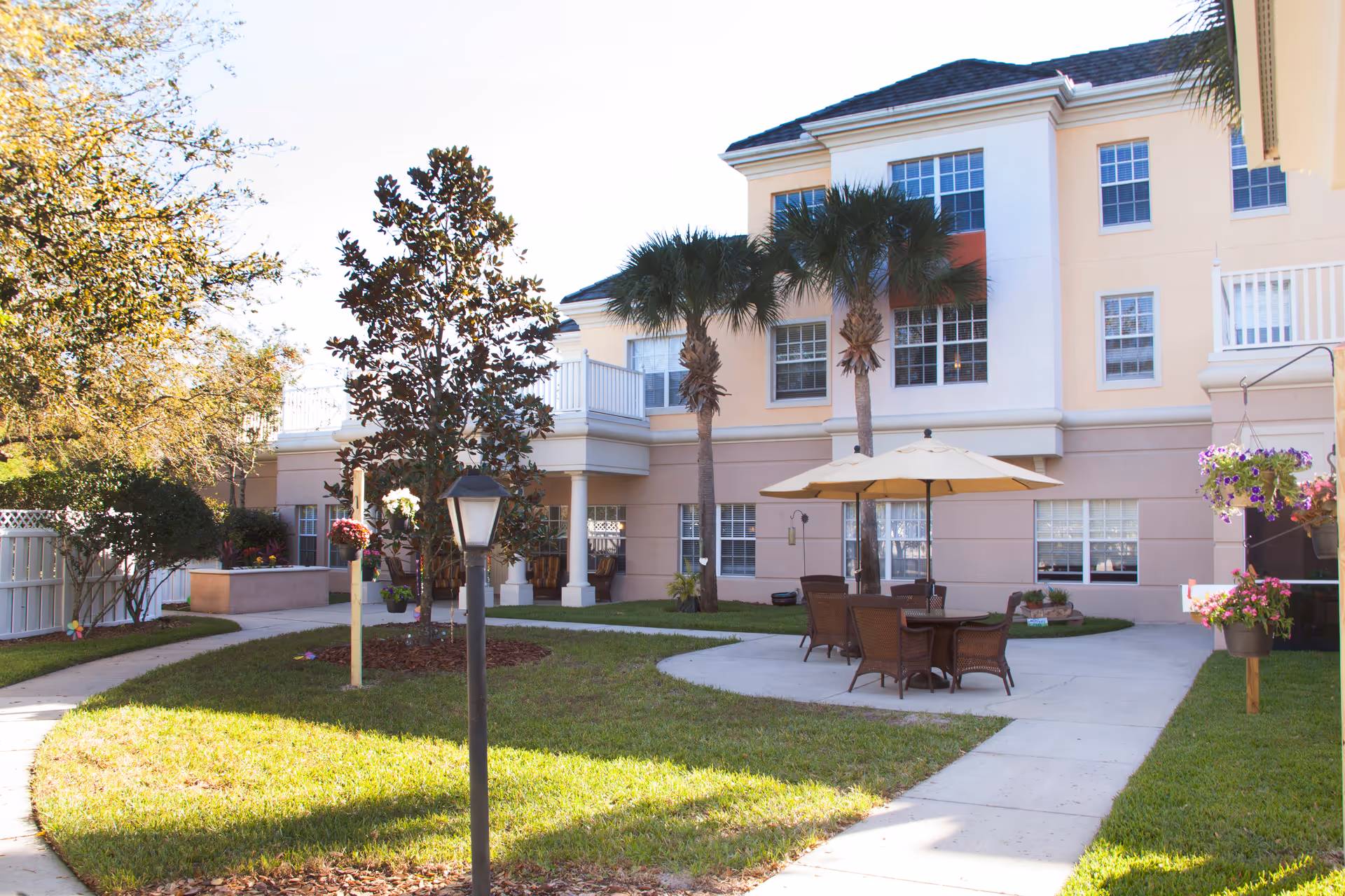 Outdoor courtyard area at Tampa Gardens Senior Living with a paved walkway, green grass, palm trees, a table with chairs under a large umbrella, and hanging flower pots near a multi-story building.