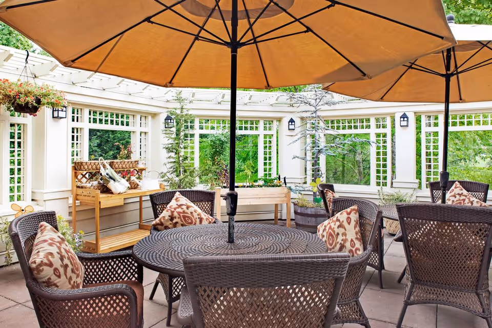 Outdoor patio area with round wicker tables and chairs, each chair having patterned cushions. Large beige umbrellas provide shade. The patio is surrounded by white lattice walls with windows and hanging flower baskets, with greenery visible outside.