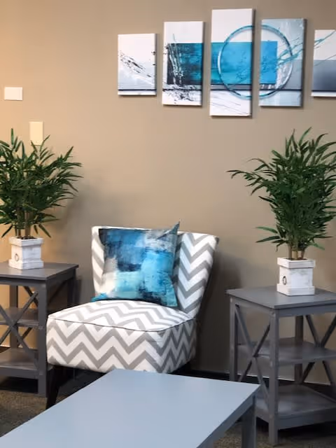 A cozy seating area with a chevron-patterned armchair featuring a blue and white abstract cushion, flanked by two gray side tables each holding a potted green plant. Above the chair is a five-panel abstract wall art in shades of blue and white. A light gray coffee table is in the foreground.