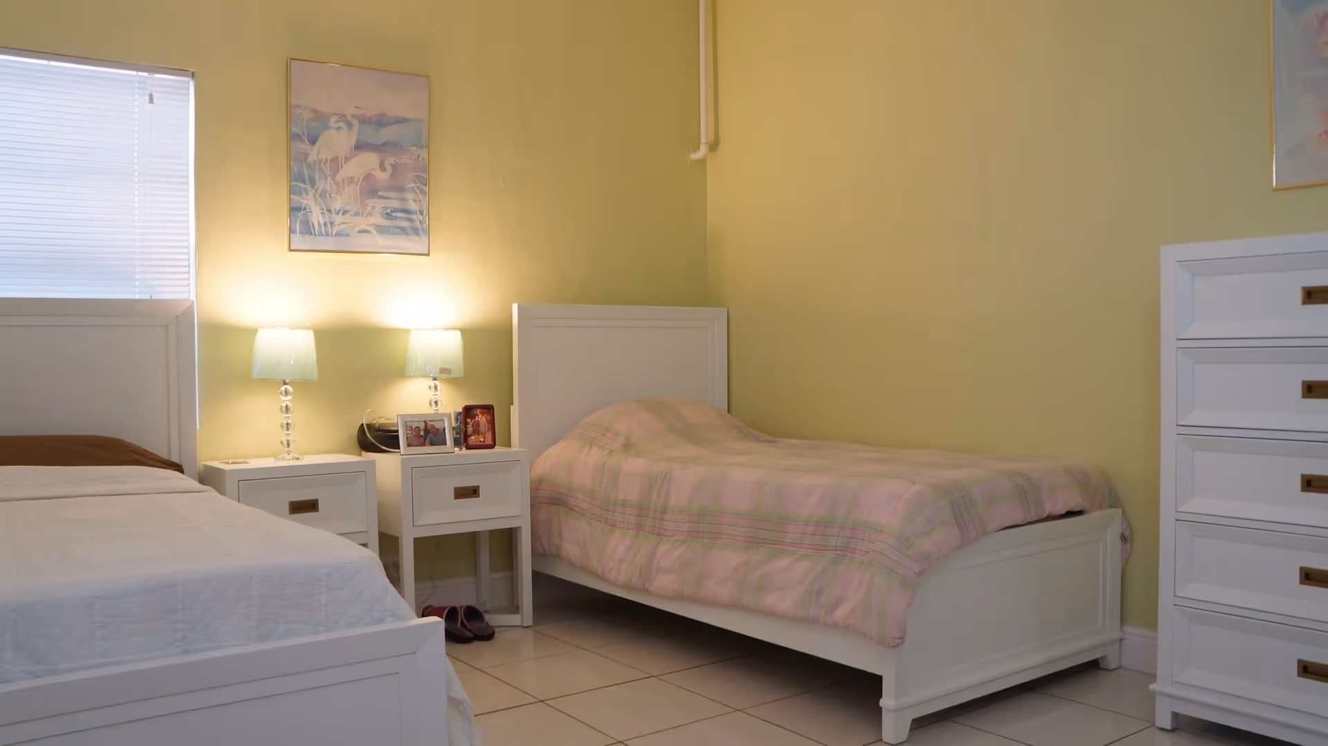 Two twin beds in a softly lit bedroom with nightstands, lamps, framed art, and a white dresser against pale yellow walls.