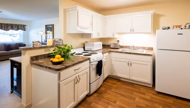 A bright kitchen with white cabinets, a white stove, microwave, toaster, and refrigerator. The countertops are brown with a bowl of lemons and a green plant on one side. The kitchen opens into a living room area with a couch and window with a valance.