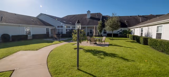 Sunlit courtyard with a curved walkway, green lawn, patio seating, lamp post, and surrounding single-story senior living buildings.