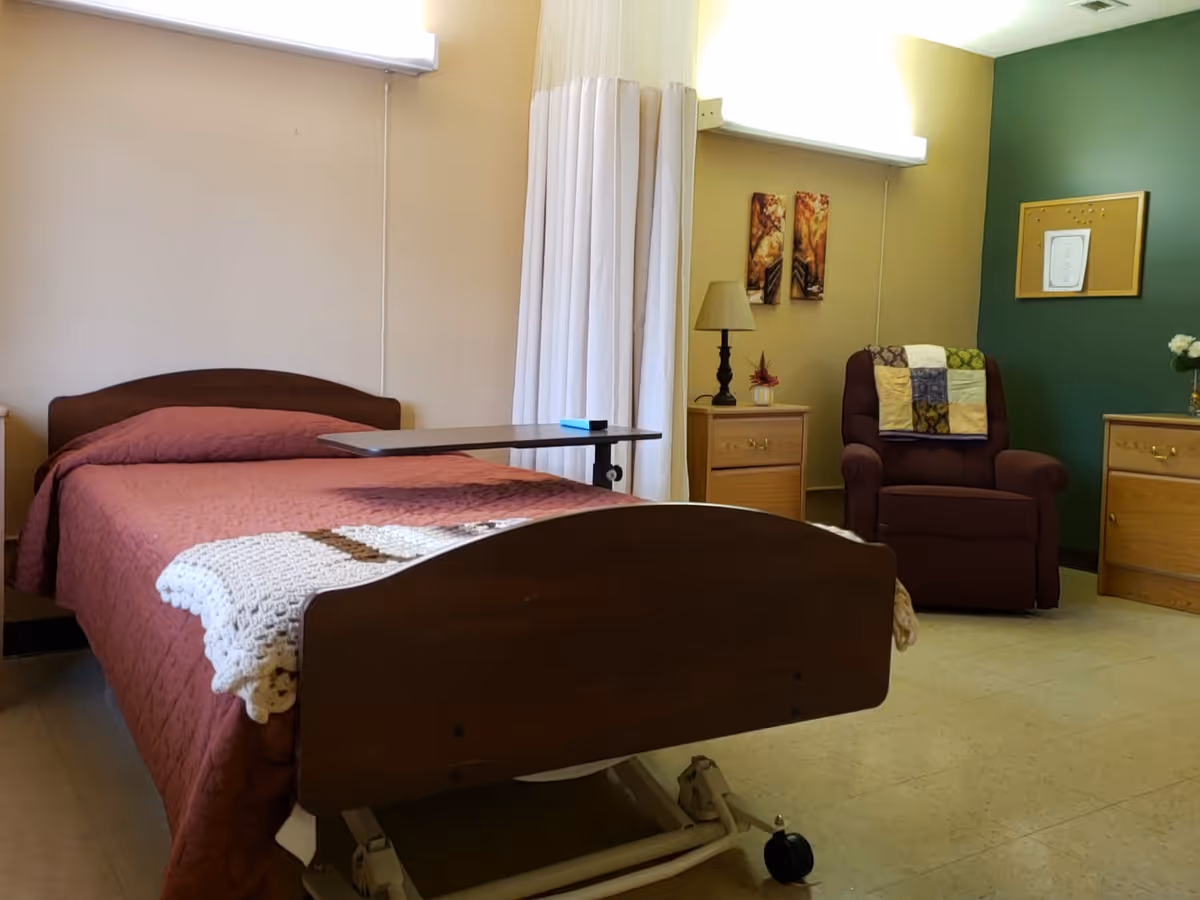 A senior living facility bedroom with a hospital-style bed covered in a red quilt and a white crocheted blanket at the foot. There is a wooden bedside table with a lamp and a small plant, a brown recliner chair with a patchwork quilt draped over the back, and two framed pictures on the wall. The walls are painted beige and green, and there is a window with white curtains.
