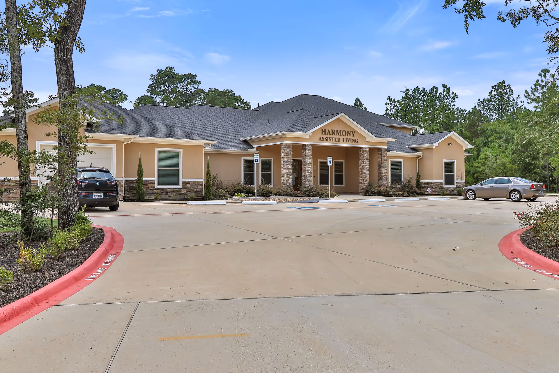 Exterior front view of Harmony Assisted Living facility with a beige building featuring stone accents, a covered entrance with columns, a parking lot with two cars, and surrounding trees under a blue sky.