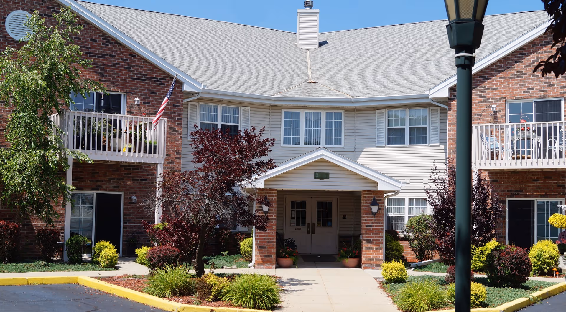 Front entrance of a two-story brick-and-siding retirement apartment building with balconies, landscaping, and a walkway.