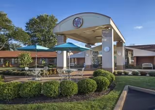 Covered entrance and porte-cochère of a senior living facility with patio tables, umbrellas, and manicured landscaping.