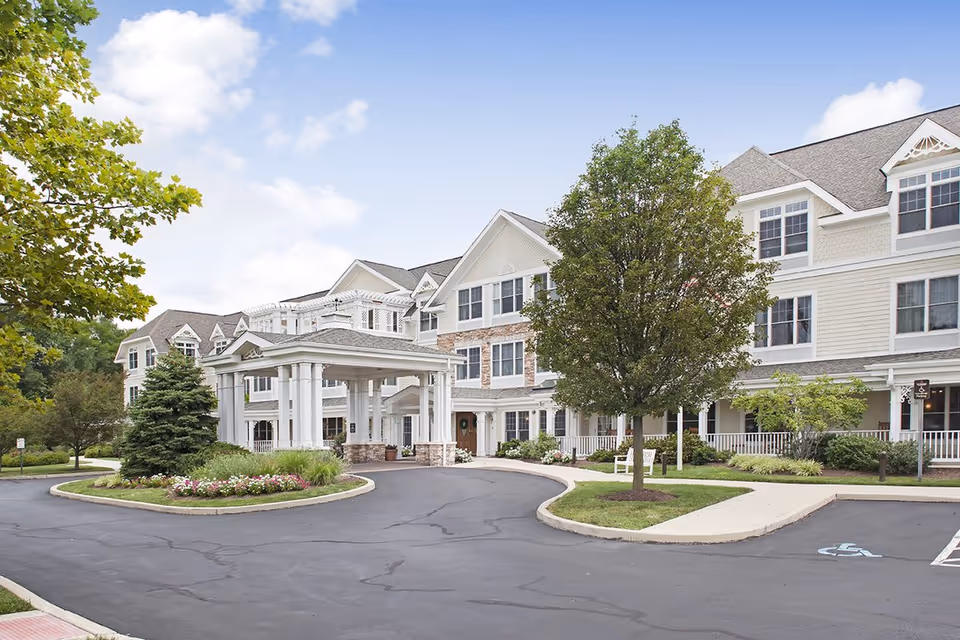 Exterior view of a large, multi-story senior living facility with white siding and stone accents. The building has a covered entrance with columns, surrounded by landscaped greenery, trees, and flower beds. The sky is partly cloudy.
