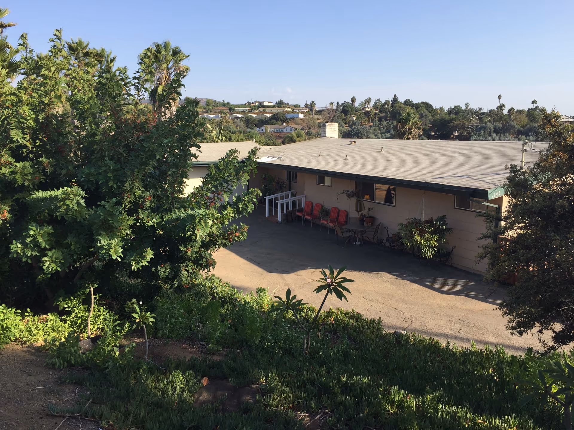 Exterior view of a single-story building with a flat roof surrounded by greenery and trees. There are several red chairs lined up against the building wall on a paved area. The sky is clear and blue.