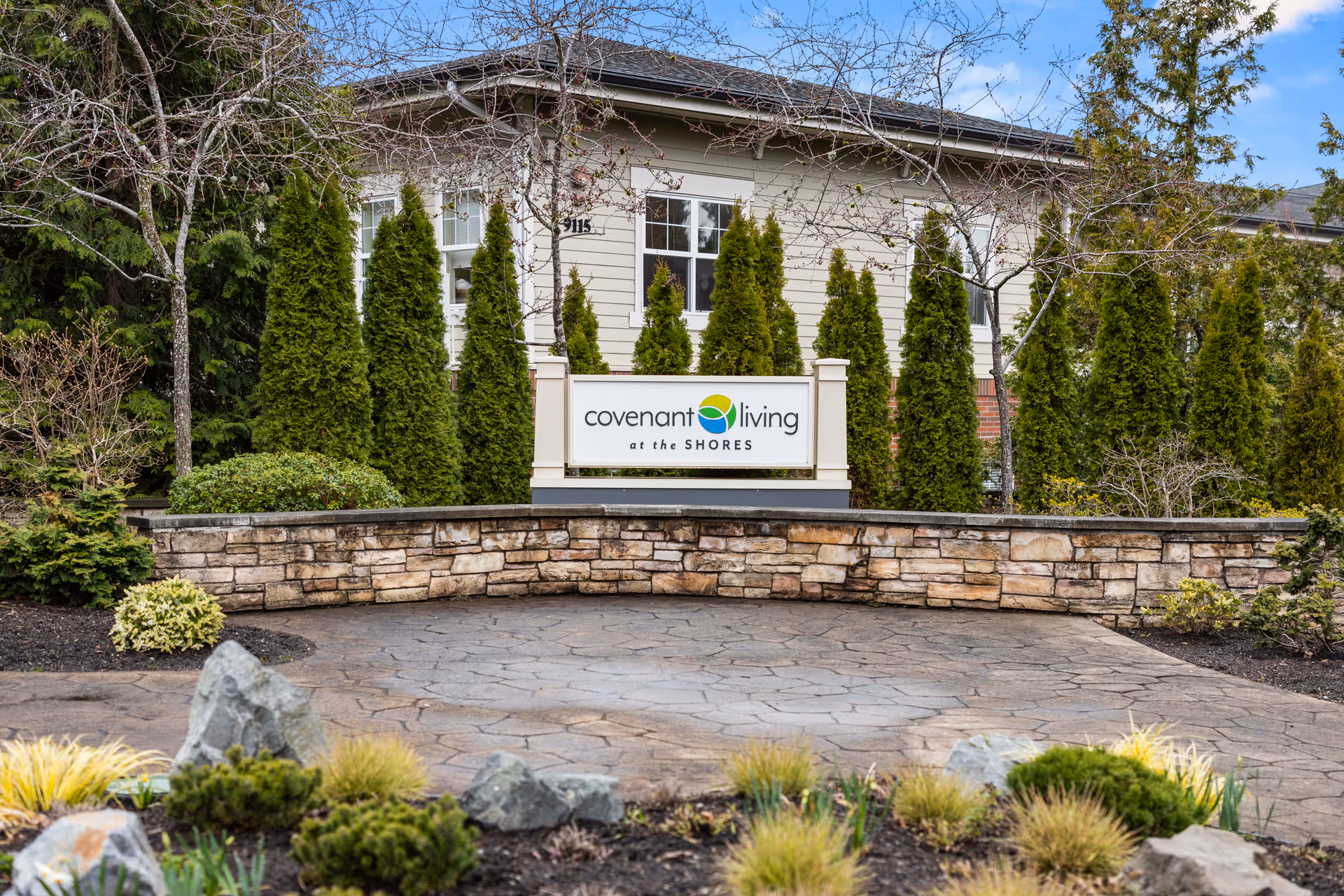 Outdoor view of Covenant Living at the Shores facility sign surrounded by tall green shrubs and trees, with a stone wall and landscaped garden in the foreground.