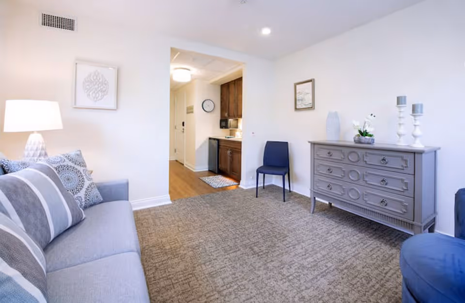 A bright living room with a gray sofa adorned with patterned pillows on the left, a lamp on a side table, a blue chair against the far wall, and a gray dresser with decorative items on the right. The room has beige carpeting and white walls with framed artwork. An open doorway leads to a small kitchen area with wooden cabinets and a clock on the wall.