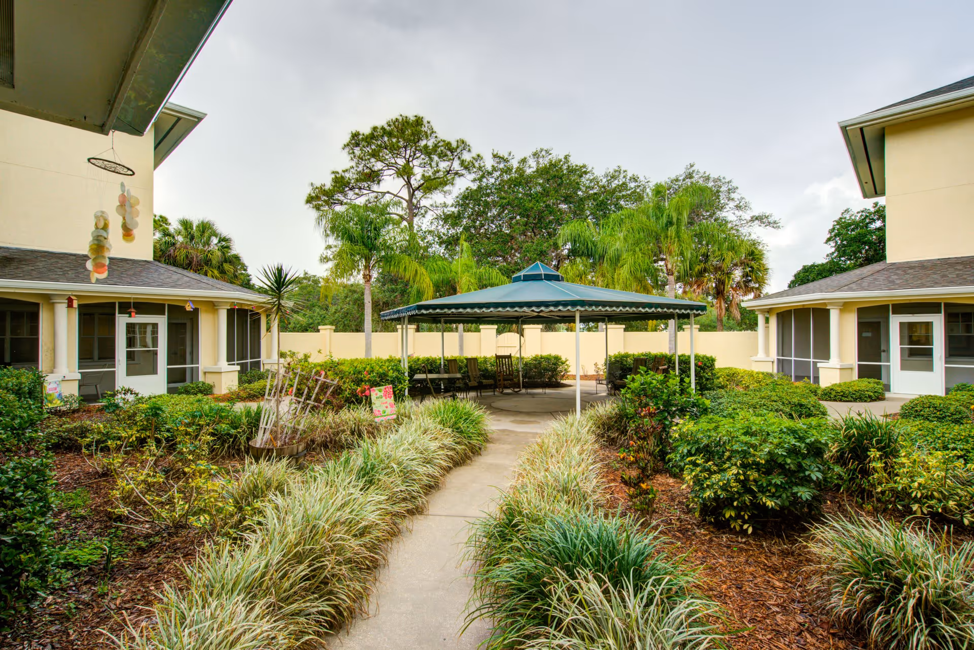 Outdoor courtyard area with a paved walkway leading to a covered seating area with chairs. The courtyard is surrounded by green bushes, plants, and trees, with two beige buildings on either side featuring screened doors and windows.