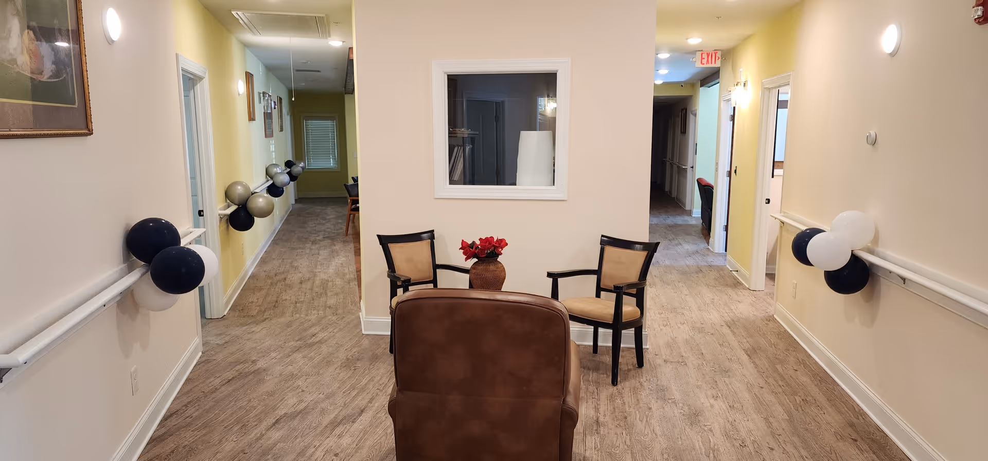 A hallway in a senior care facility with light-colored walls and wood flooring. There are handrails on both sides of the hallway decorated with black and white balloons. At the end of the hallway, there are three chairs arranged around a small table with a vase of red flowers. The hallway leads to other rooms and has ceiling lights and an exit sign visible in the background.