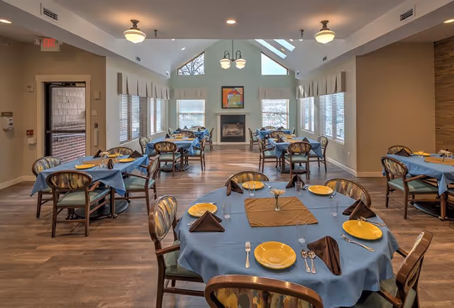 A dining room in a senior living facility with multiple round and square tables covered with blue tablecloths, set with yellow plates, brown napkins, silverware, and glasses. The room has large windows on both sides, a fireplace at the far end, wooden flooring, and ceiling lights.