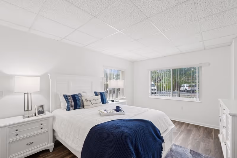 Bright and clean bedroom with a white bed frame, white bedding, and blue accent pillows and blanket. Two windows with vertical blinds let in natural light. There are two white nightstands with lamps and a framed photo on one side, and a white dresser on the right side of the room. The floor is wood with a small rug partially visible.