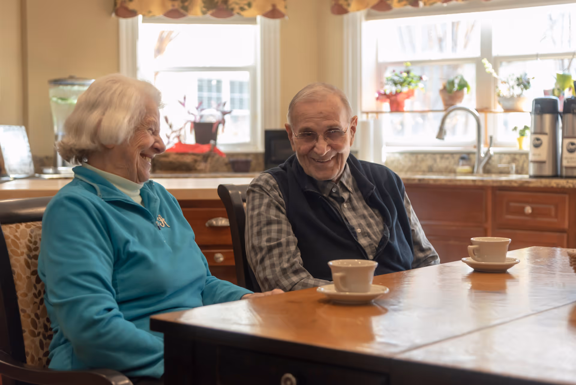 An elderly woman and an elderly man sitting at a wooden table in a kitchen or dining area, smiling and enjoying each other's company. There are two cups on the table, and the background shows kitchen cabinets, a sink, and windows with potted plants on the sill.