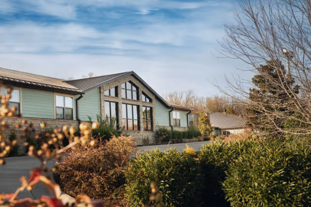 Exterior view of a single-story assisted living facility building with large windows, surrounded by bushes and trees under a partly cloudy sky.