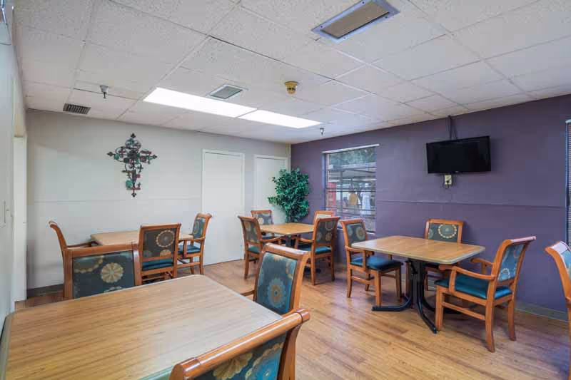 Interior view of a dining room in a senior living facility with several wooden tables and chairs with patterned upholstery. The room has a wooden floor, a purple accent wall with a mounted flat-screen TV, a window, and a decorative wall hanging on a white wall. There is also a green potted plant in the corner.