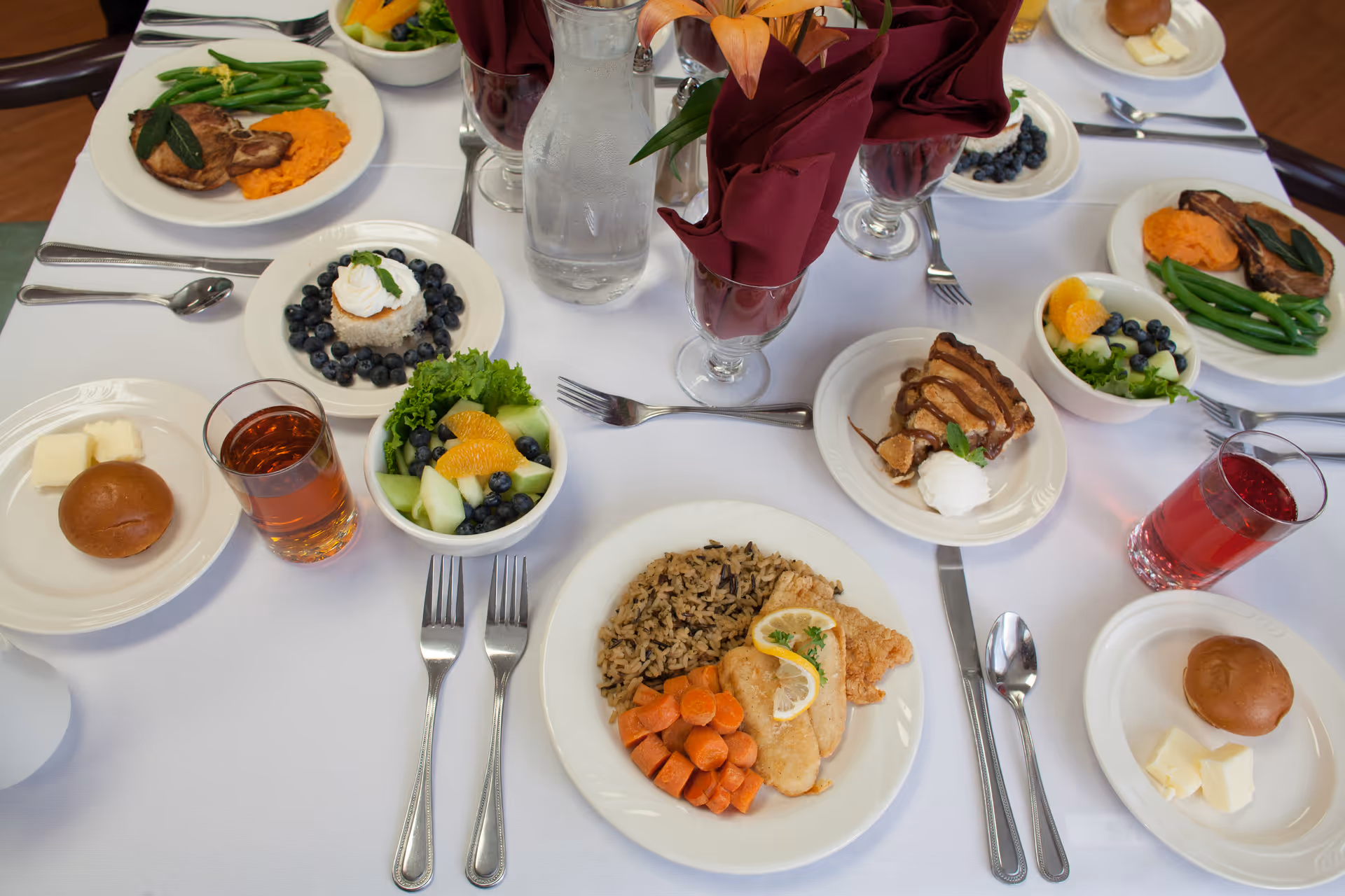 A dining table set with plates of food including grilled chicken with lemon slices, wild rice, cooked carrots, green beans, mashed sweet potatoes, fresh fruit salad, blueberry dessert with whipped cream, bread rolls with butter, and glasses of iced tea and red juice. The table is covered with a white tablecloth and has burgundy cloth napkins in glasses with a flower centerpiece.