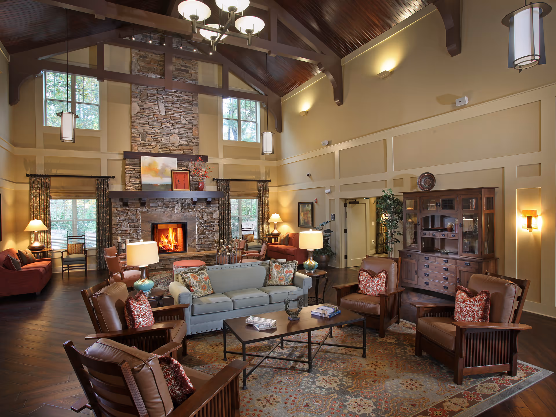 A spacious and warmly lit living room with high vaulted wooden ceilings and large windows. The room features a stone fireplace with a fire burning, surrounded by comfortable seating including a light blue sofa, several brown leather armchairs with red patterned pillows, and red couches. There are multiple lamps providing soft lighting, a large patterned rug, and a wooden cabinet against one wall. The room has a cozy and inviting atmosphere.