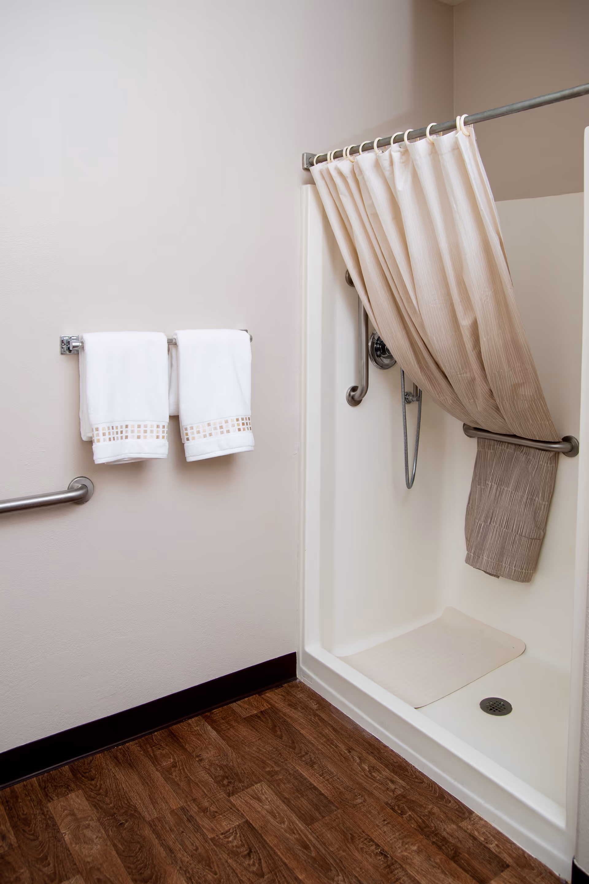 A clean shower area with a beige shower curtain partially pulled to the side, a white shower base with a non-slip mat, and stainless steel grab bars. Two white towels with decorative trim hang on a towel rack on the adjacent wall. The floor is wood-patterned.