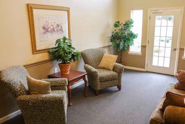 A cozy sitting area with two patterned armchairs and a brown sofa, each with cushions. A wooden side table between the armchairs holds a potted plant. There is a framed artwork on the wall above the table. The room has beige walls, a carpeted floor, and a door with windows letting in natural light.