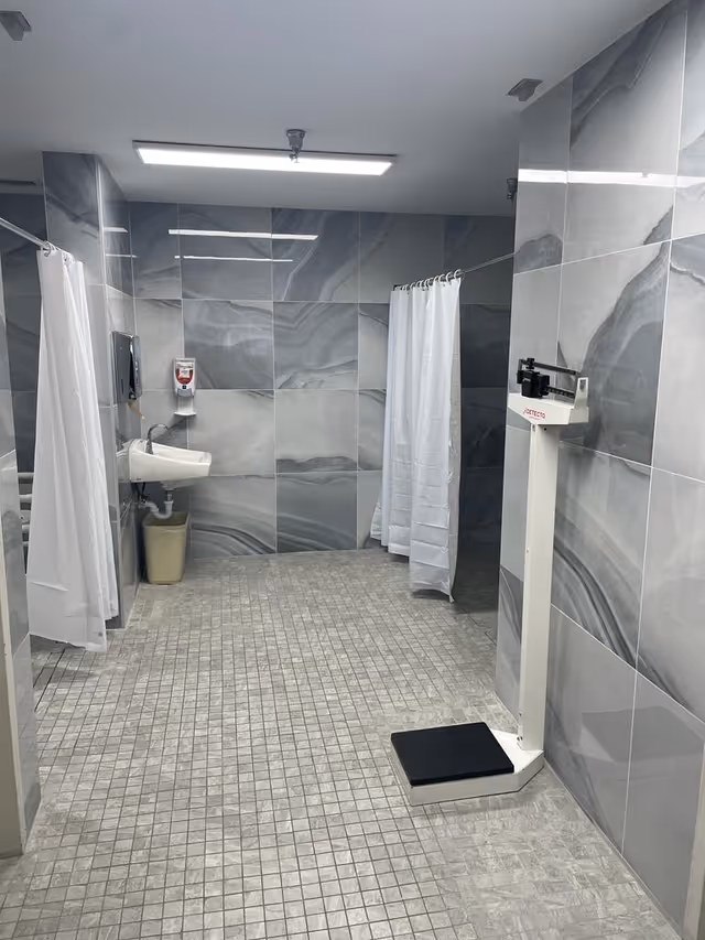 A clean, modern bathroom with gray marble-patterned tiled walls and small square tiled floor. The room features two shower stalls with white curtains, a white wall-mounted sink with a soap dispenser above it, a beige trash bin underneath, and a white mechanical weight scale with a black platform on the right side.