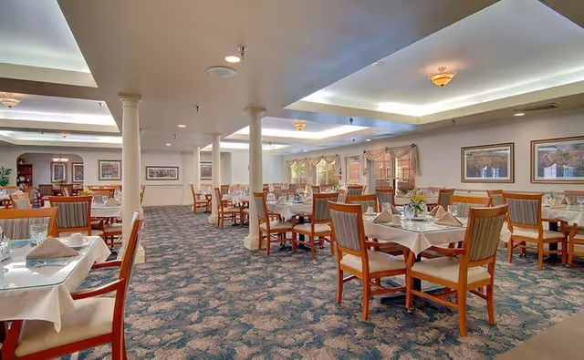 A spacious dining room in a senior living facility with multiple tables covered with white tablecloths, each set with glassware, napkins, and silverware. The room has carpeted floors, several columns, framed artwork on the walls, and large windows with valances allowing natural light to enter.