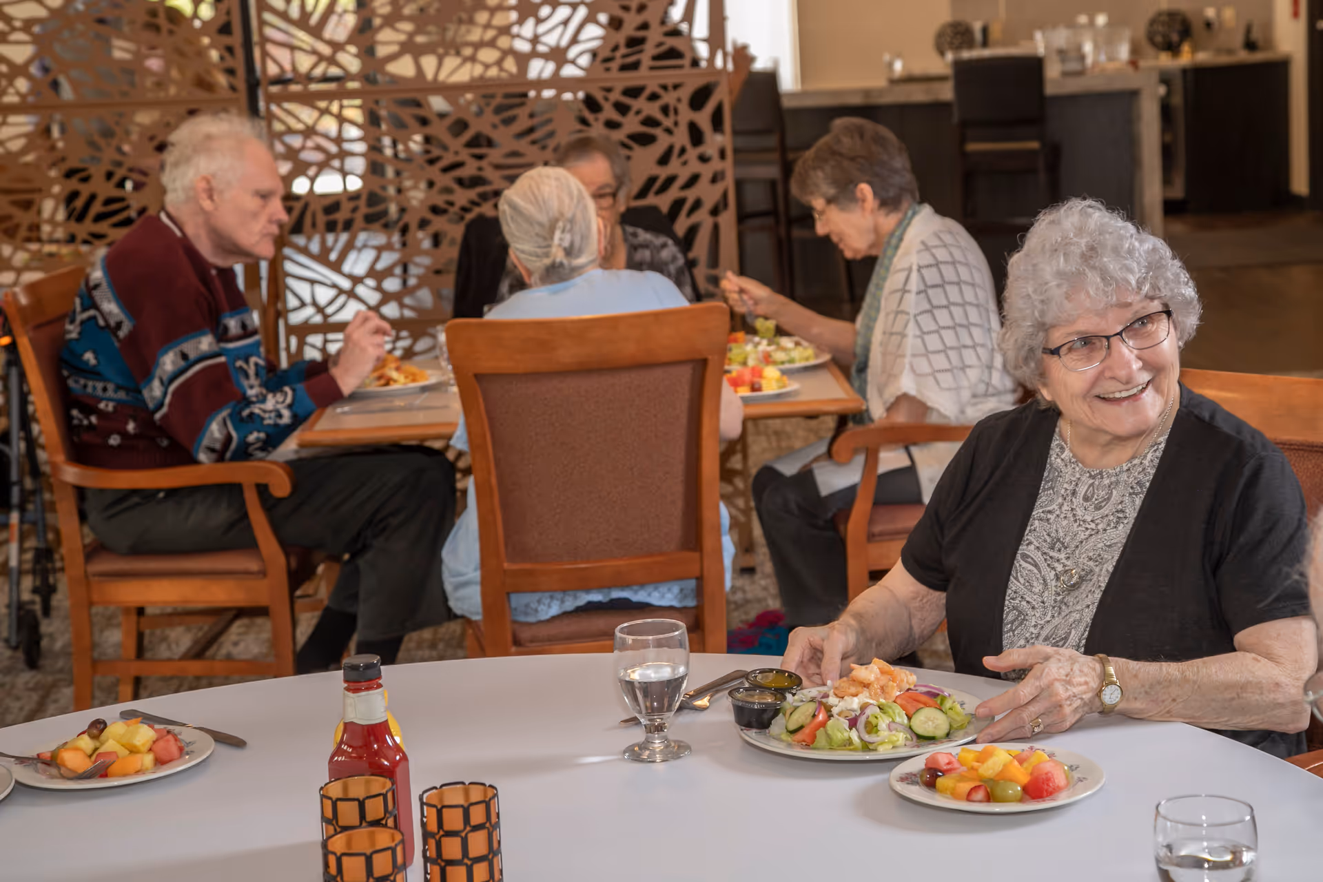 Several elderly residents dining together in a communal dining room, with a smiling woman in the foreground and plates of salad and fruit on the table.