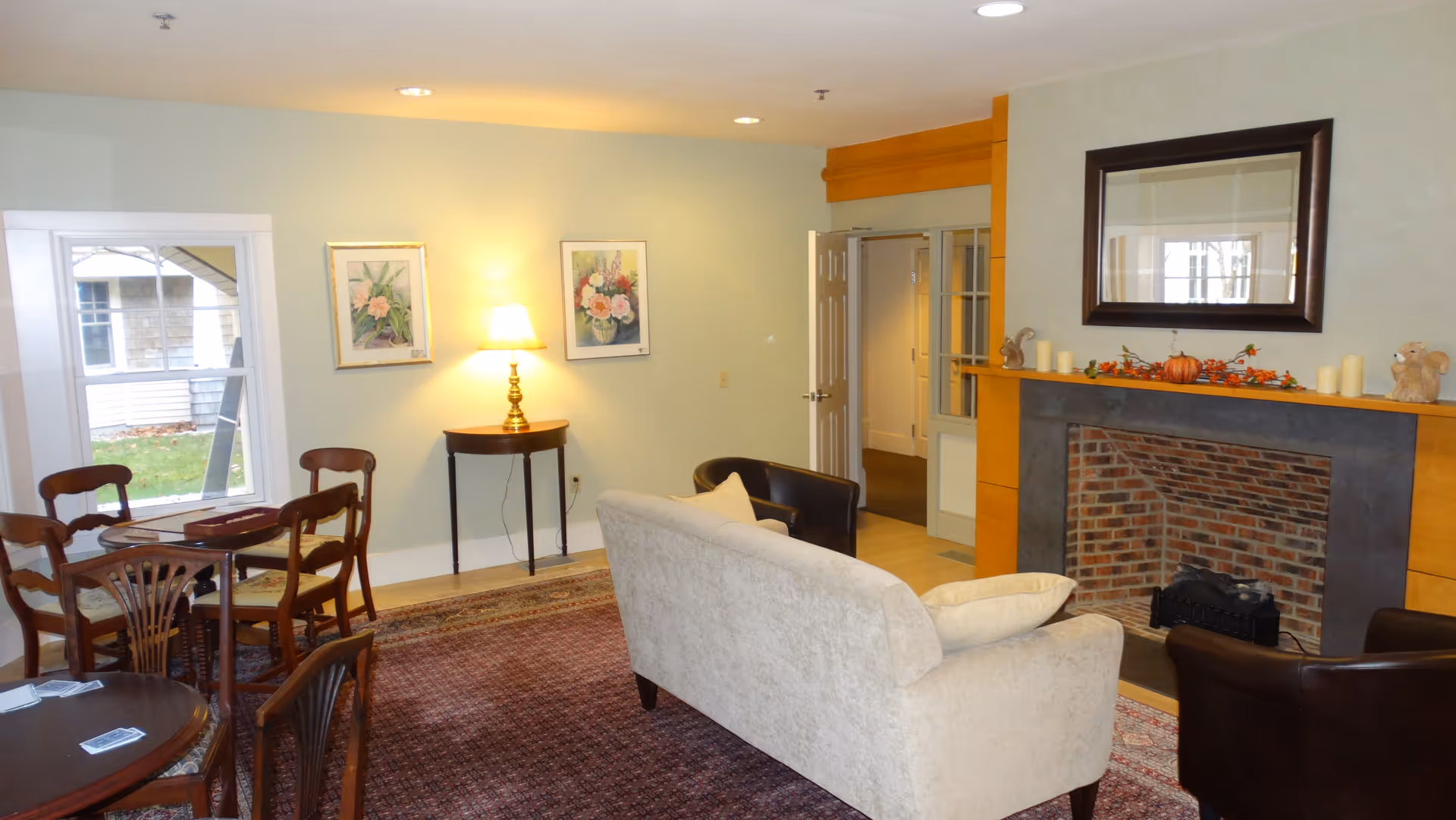 A cozy common living room with a sofa and armchairs facing a fireplace, a small dining table, and wall art illuminated by a table lamp.