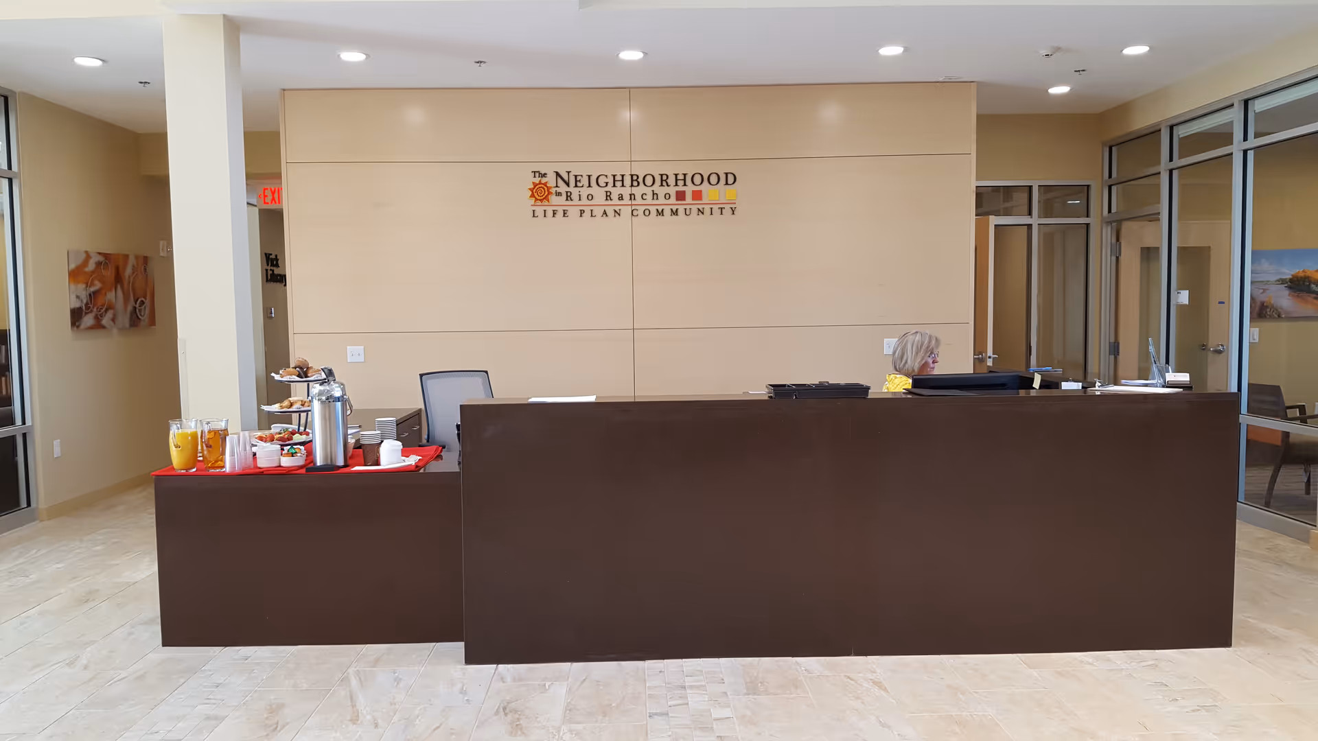 Reception area of The Neighborhood in Rio Rancho Life Plan Community with a dark brown front desk. A woman is seated behind the desk working on a computer. On the left side of the desk, there is a table with refreshments including juice, coffee, cups, and snacks. The background features a light wood panel wall with the community's logo and name displayed. Glass doors and windows are visible on the right side.