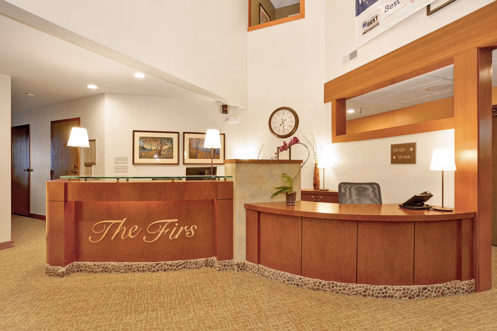Reception area of The Firs facility featuring a wooden front desk with the name 'The Firs' engraved on it, two table lamps, a clock on the wall, framed artwork, and directional signage for rooms 101-120 and 121-144.