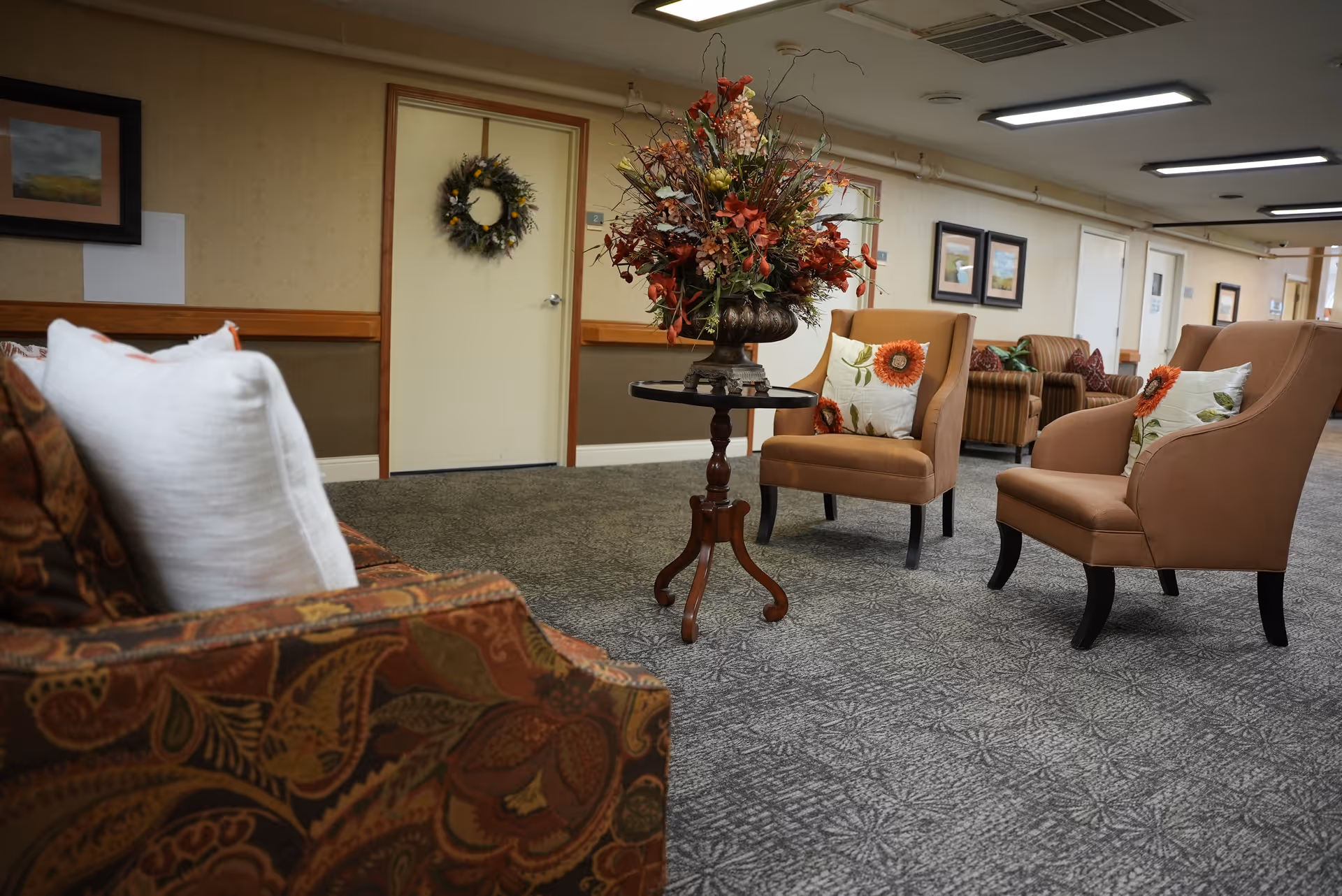 A senior living facility seating area with upholstered chairs, a small round table topped with a floral arrangement, and framed art on the walls.