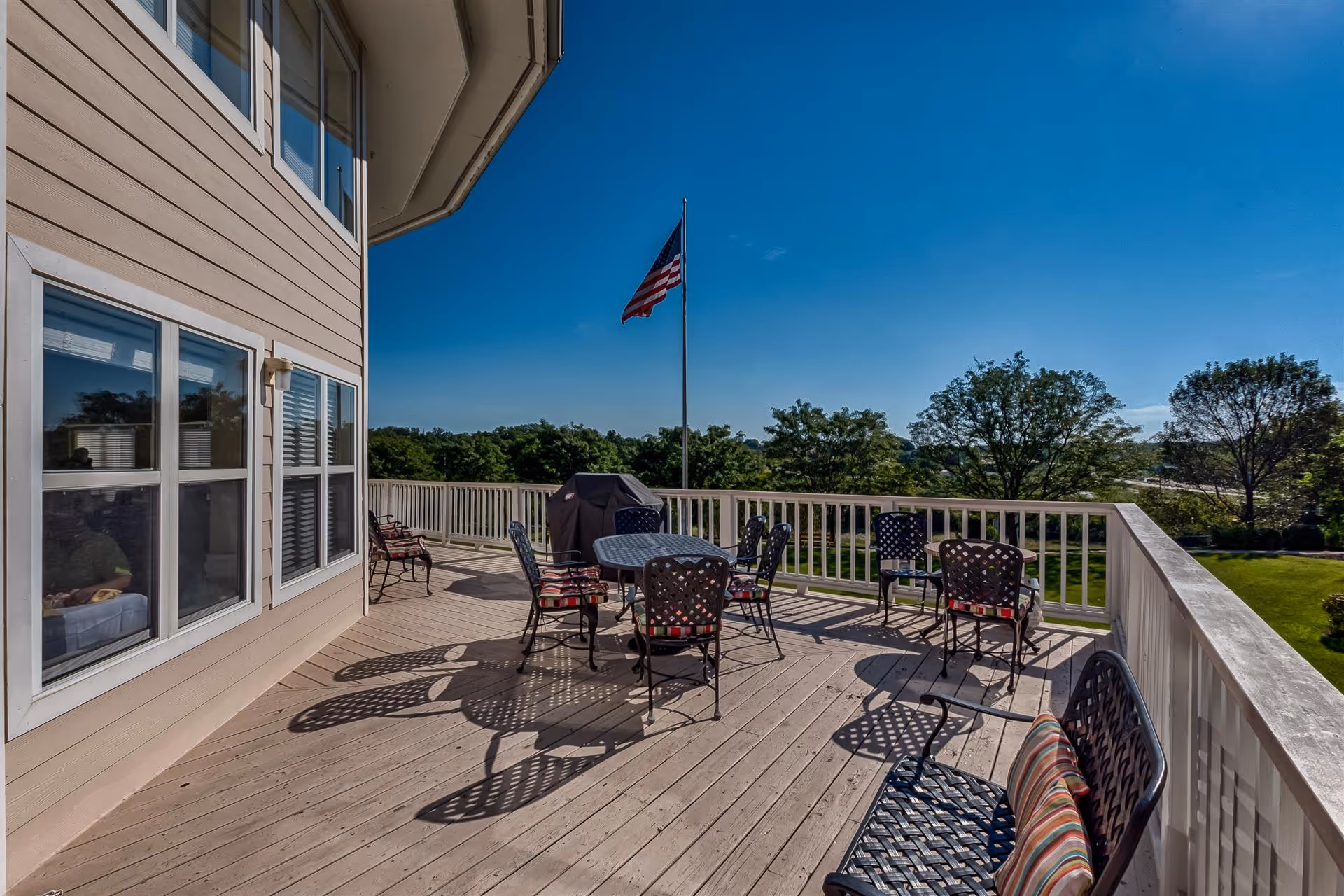 Outdoor wooden deck area with metal chairs and tables, some with cushions, a grill covered with a protective cover, an American flag on a flagpole, and a view of trees and a clear blue sky.