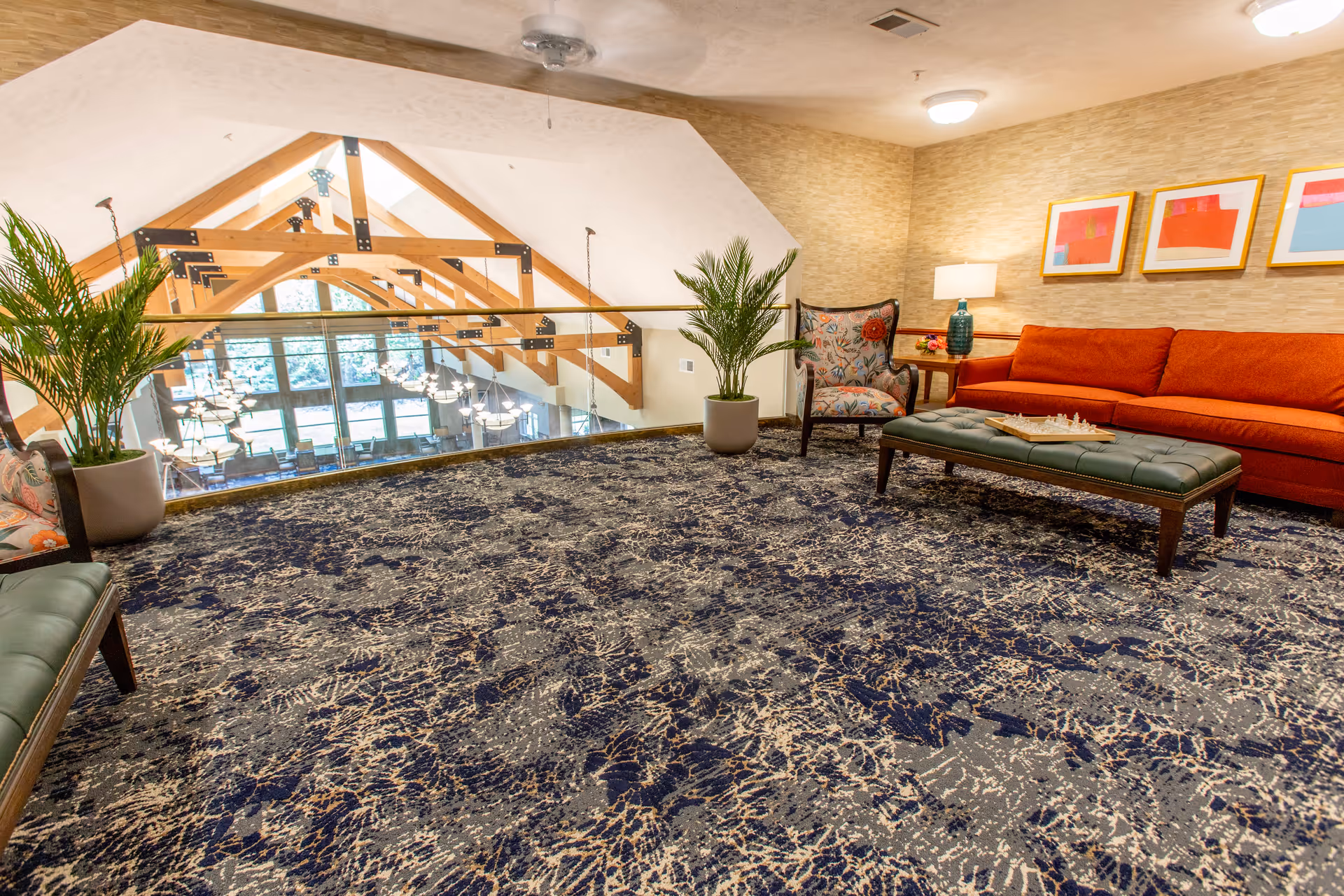A cozy indoor lounge area with a patterned carpet, an orange sofa, a floral armchair, a green cushioned bench with a chessboard on top, two potted plants, a side table with a lamp, and three colorful framed artworks on the wall. The space overlooks a large room below with wooden beams and multiple hanging lights.