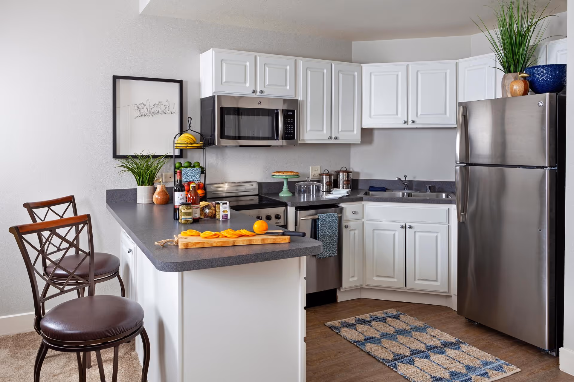 Modern kitchen with white cabinets, stainless steel refrigerator, microwave, and dishwasher. A countertop with two brown cushioned bar stools, a cutting board with sliced mango, various condiments, and decorative plants are visible. A framed artwork hangs on the wall, and a patterned rug is on the floor.