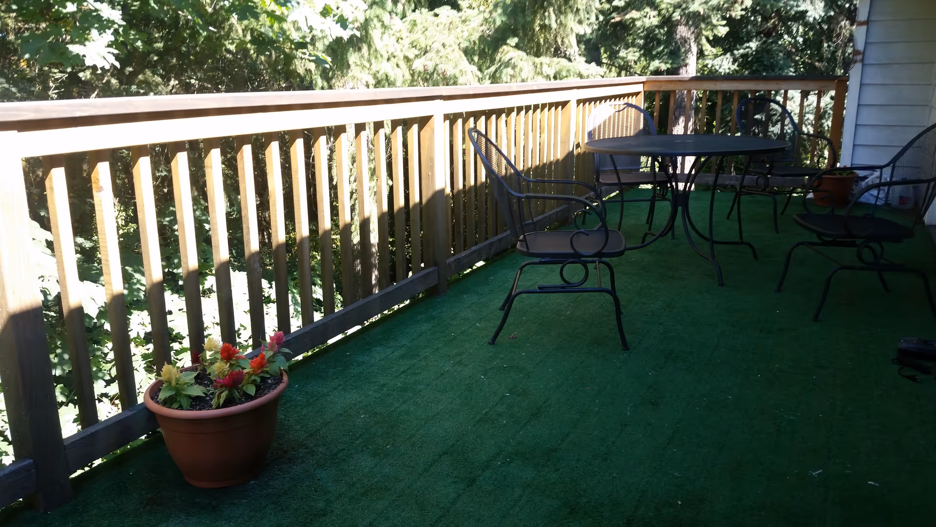 Outdoor patio area with green artificial grass flooring, a wooden railing, a round metal table, four metal chairs, and two potted plants with flowers.
