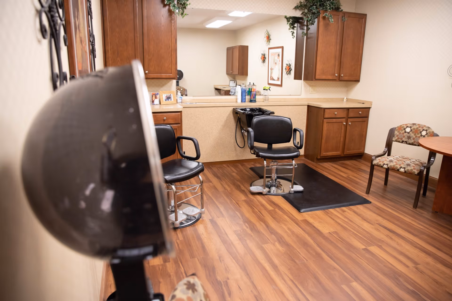 Interior view of a salon area in a senior living facility with two black salon chairs, a hair washing station, wooden cabinets, a round table with a floral-patterned chair, and a hair dryer in the foreground.