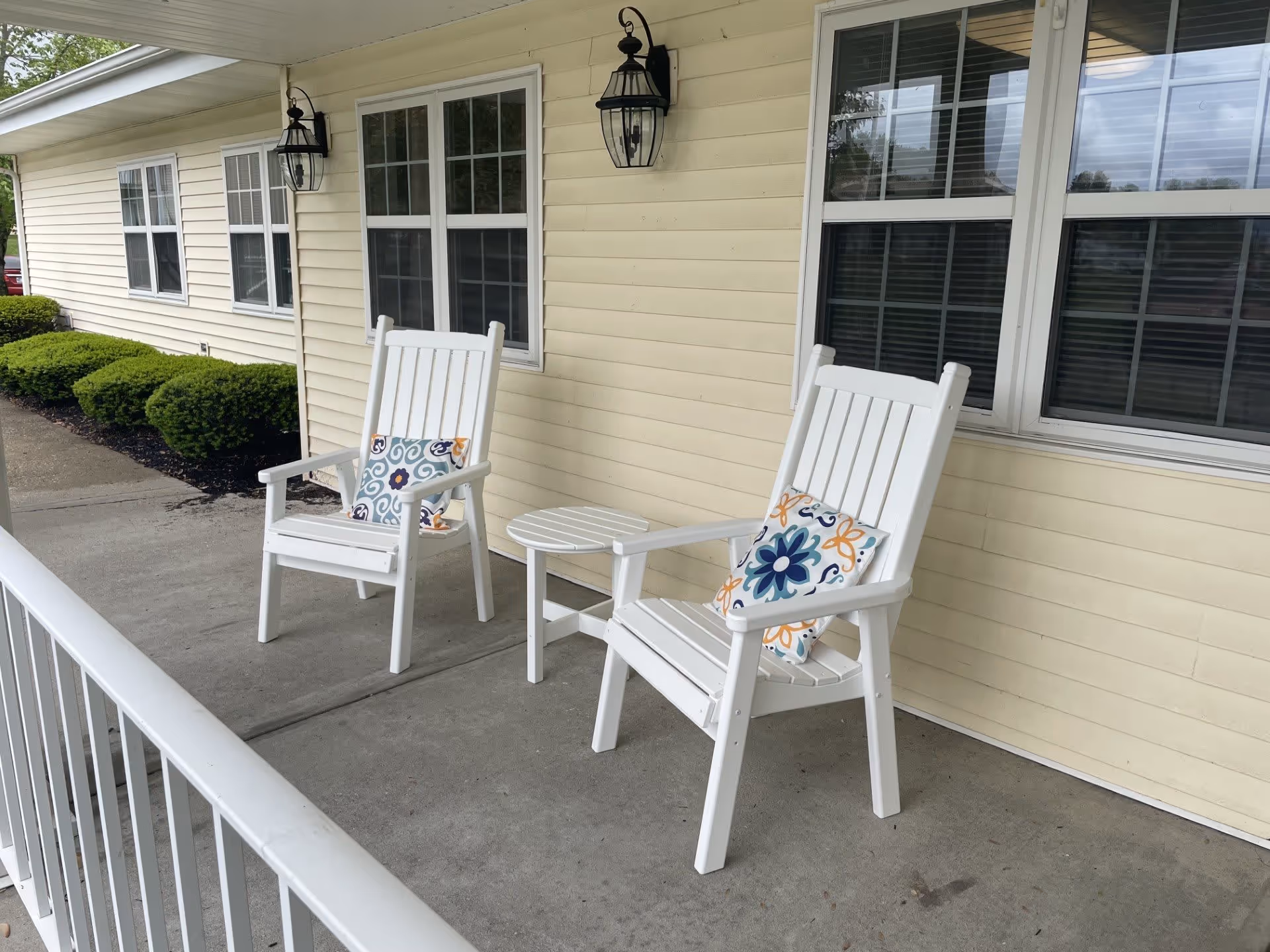 A porch area with two white wooden chairs, each with a decorative cushion featuring a floral pattern in blue, orange, and white. Between the chairs is a small white round table. The porch has a concrete floor and is attached to a light yellow building with several windows and two black outdoor wall lanterns.