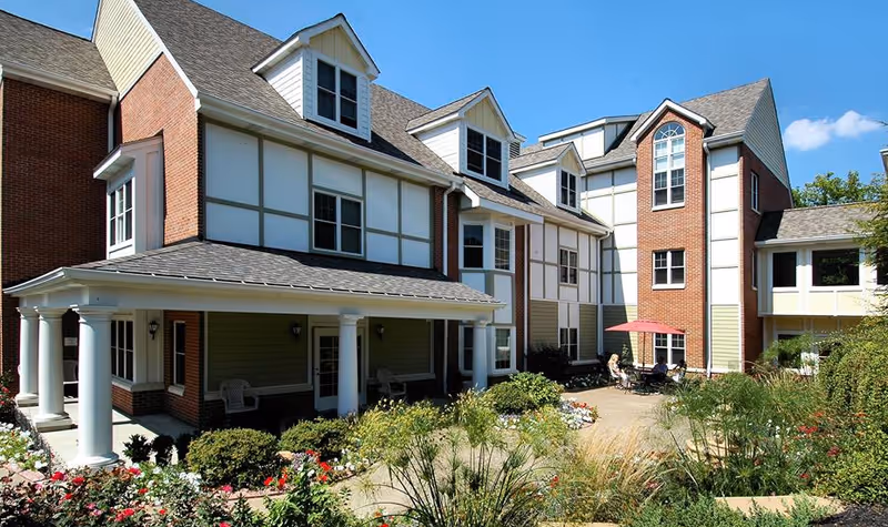 Exterior view of a multi-story senior living facility with brick and white paneling, featuring a covered porch with white columns, landscaped garden with flowers and shrubs, and a patio area with people sitting under a red umbrella.