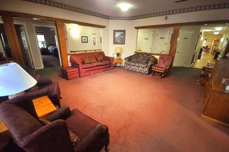 A spacious living room area in a senior living facility with a reddish carpet, multiple upholstered chairs and sofas, wooden side tables, a lamp, and framed artwork on the walls. The room has doorways leading to other rooms and a hallway with additional seating and furniture visible in the distance.