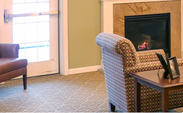 Cozy seating area with patterned armchairs, a side table with picture frames, a lit fireplace, and a glass door.