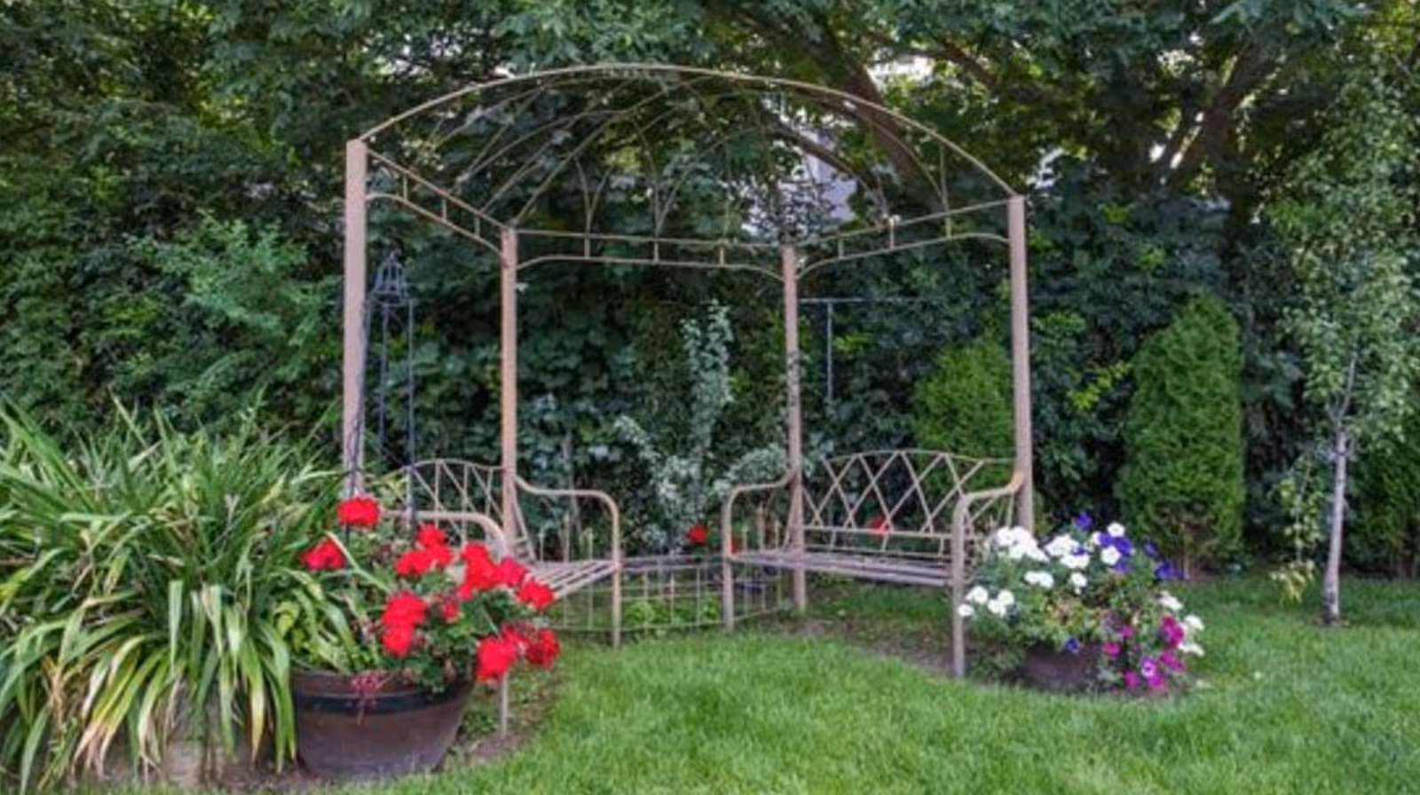 A garden area with a metal gazebo structure featuring two benches facing each other. The surrounding area has green grass, various plants, and colorful flowers including red, white, and purple blooms. Trees and dense greenery form the background.