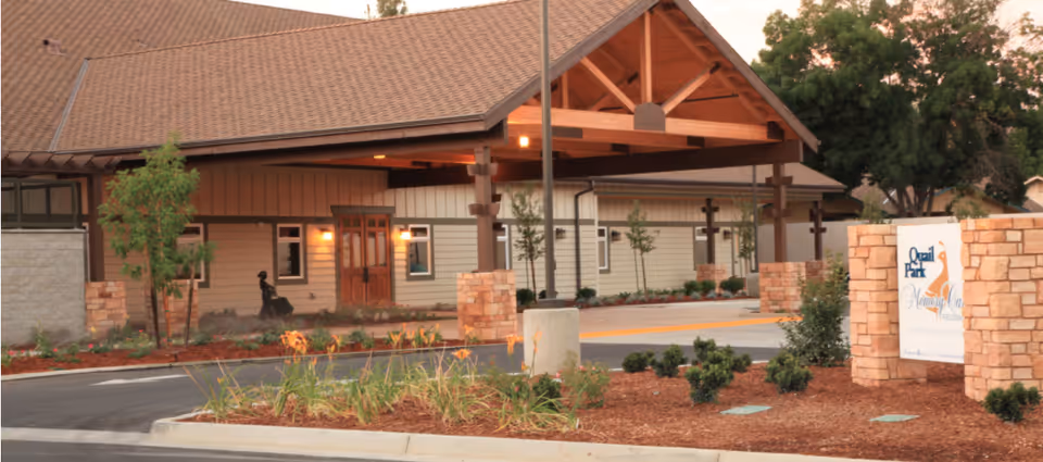 Entrance view of Quail Park Memory Care Residences of Visalia showing a covered drop-off area with wooden beams, a landscaped garden with plants and mulch, and a stone sign with the facility's name.