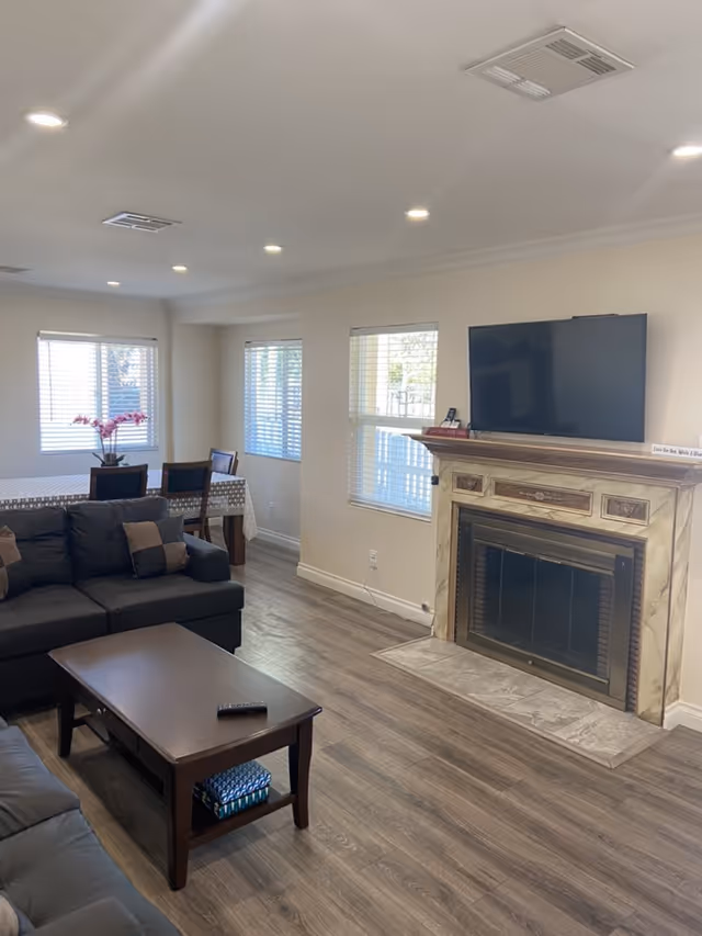 Well-lit living room with sofas, a coffee table, dining area in the background, and a fireplace with a TV mounted above it.