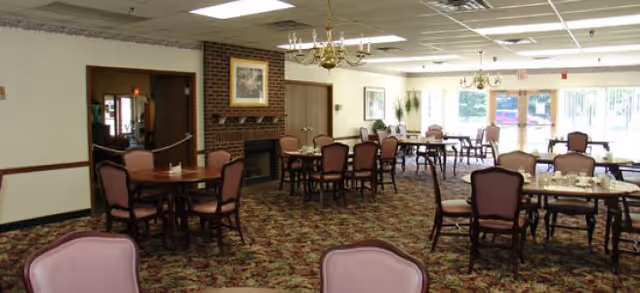 Carpeted dining room with round tables, pink-upholstered chairs, chandeliers, and a brick fireplace.