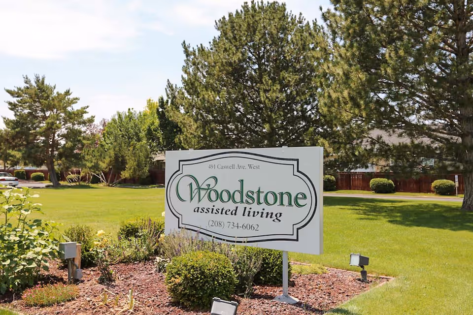 A Woodstone Assisted Living sign on a landscaped lawn with trees and residential buildings in the background.