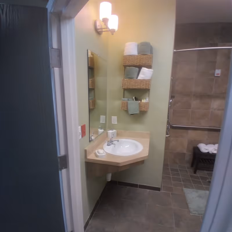 View into a bathroom with a wall-mounted sink and mirror. Above the sink are three wicker baskets holding folded towels and toiletries. The bathroom has tiled floors and walls, with a walk-in shower area visible in the background, equipped with grab bars and a small bench with rolled towels.