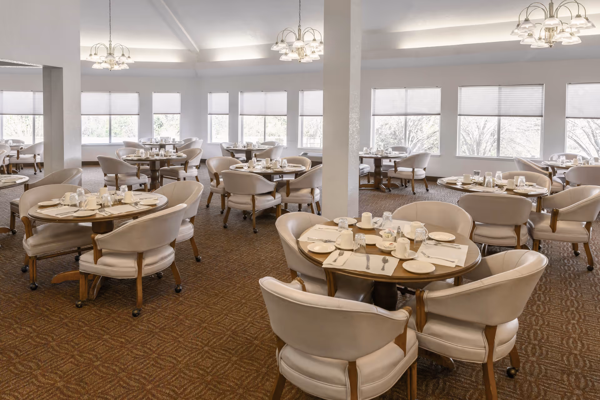 A spacious dining room with multiple round tables set for meals, each table surrounded by cushioned chairs. The room has large windows letting in natural light and chandeliers hanging from the ceiling. The carpet has a patterned design and the walls are painted white.